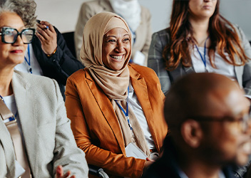 A group of people seated in an auditorium, all appearing attentive and engaged. Some individuals are wearing lanyards, suggesting a conference setting. The background is blurred, highlighting the focus on the attendees.